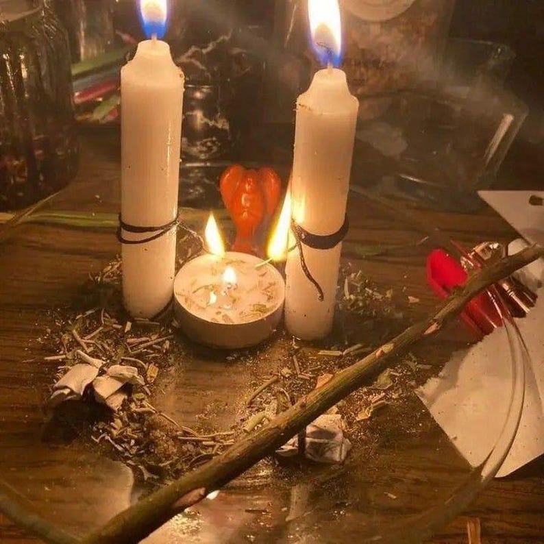 Puede incluir: Una disposici&oacute;n espiritual con dos velas blancas altas, una peque&ntilde;a vela de t&eacute; encendida y una decoraci&oacute;n en forma de coraz&oacute;n rojo. Tambi&eacute;n hay un palo de madera y hierbas secas, creando un ambiente m&iacute;stico.