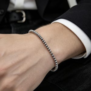 May include: A silver-toned beaded bracelet featuring a continuous row of small, uniform, metallic spheres. The beads have a polished finish with darkened detailing between each individual bead, creating a textured, linked appearance on the wrist.