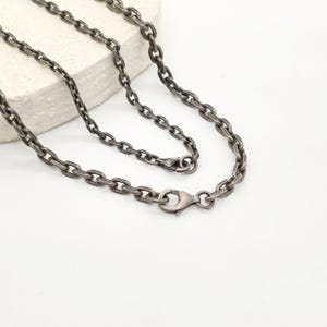 May include: A dark oxidized silver chain necklace featuring oval-shaped links with a textured, matte finish. The necklace is secured with a lobster claw clasp, showcasing a consistent link pattern throughout the entire length of the chain.