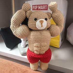 Puede incluir: Un oso de peluche de ganchillo con brazos y torso musculosos, que lleva pantalones cortos rojos. El oso tiene ojos negros y un hocico blanco y marrón. El fondo incluye una bolsa blanca con "POP MART" impreso en rojo.