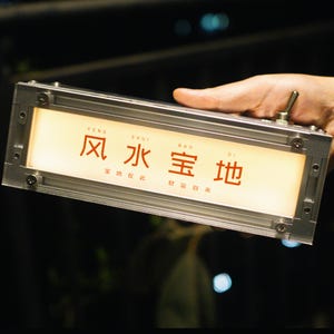 May include: A rectangular, illuminated sign with a silver frame. The sign displays Chinese characters in red against a warm, glowing background. The sign is held by a hand, with a toggle switch visible.