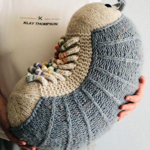 Könnte beinhalten: Ein großes, handgestricktes Gürteltier-Plüschtier. Das Plüschtier hat einen hellgrauen Körper mit dunkleren, gerippten Details, einen beigefarbenen Kopf und farbige Akzente. Das Gürteltier wird vor einem weißen T-Shirt mit dem Text "Klay Thompson" gehalten.