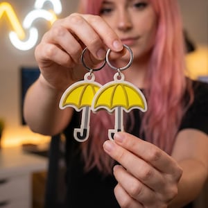 Könnte beinhalten: Zwei gelbe schirmförmige Schlüsselanhänger mit weißen Umrissen und silbernen Ringen werden gezeigt. Die Schirme haben einen schwarzen Griff. Der Hintergrund ist verschwommen, mit einem Neonzeichen und einer Person mit rosa Haaren.