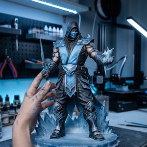 May include: A detailed figurine of a character from the Mortal Kombat video game series, Sub-Zero. The figure is predominantly blue and black, holding a sword and ice. It stands on a base of ice-like structures. The figure is approximately 10 inches tall.