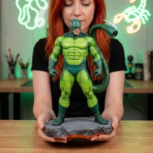 Peut inclure: Figurine d'action Scorpion vert et vert foncé. La figurine est posée sur un socle rocheux gris avec des accents orange. La figurine a une queue de scorpion et est dans une pose de super-héros.