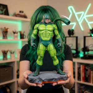 May include: A green and dark green Scorpion action figure. The figure is standing on a gray, rocky base with an orange glow. The figure has a tail with a white stinger. The figure is a collectible item.
