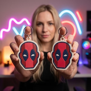 May include: Two Deadpool head-shaped keychains. Each keychain features a red and black mask design with a white border and a silver keyring. The keychains are held up in front of a blurred background.