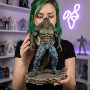 Puede incluir: Estatua detallada de una figura humanoide reptiliana, con piel verde texturizada, físico musculoso y vaqueros azules rasgados. La figura se alza sobre una base de piedra, mostrando detalles intrincados y una expresión amenazante.