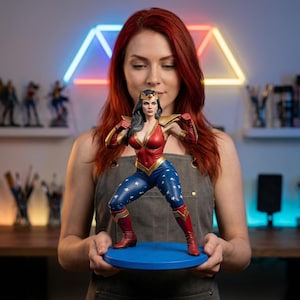 May include: A Wonder Woman statue in a fighting pose, with a red and gold bodice, blue trousers with white stars, and red boots. The statue is held on a blue circular base. The figure has a tiara and dark hair.