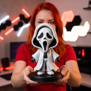 Puede incluir: Una figura estilizada del personaje Ghostface de la franquicia Scream, que presenta una túnica blanca con capucha, una máscara blanca con aberturas negras en los ojos y la boca, y guantes negros, sobre una base circular negra.