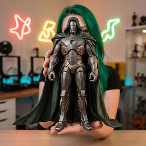 May include: A detailed action figure of a character in a metallic grey and gold armoured suit with a green cape and hood. The figure is held in front of a person with green hair. The background includes neon signs and a shelf with other figures.