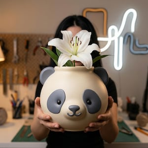 May include: A cream-colored panda-shaped ceramic vase with gray accents, holding a white lily with green leaves. The vase has a round body, gray ears, and a panda face. The vase is being held in front of a blurred background.