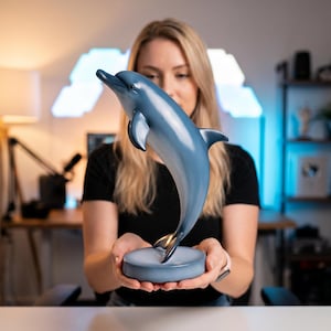 May include: A blue dolphin sculpture with a curved body and fins. The dolphin is perched on a round, gray base. The sculpture has a glossy finish and is a decorative item.