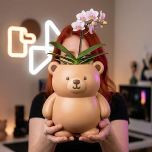 May include: A tan-colored teddy bear-shaped planter holding a flowering orchid. The planter has a smiling face and rounded features. The orchid has pink and purple flowers and green leaves.