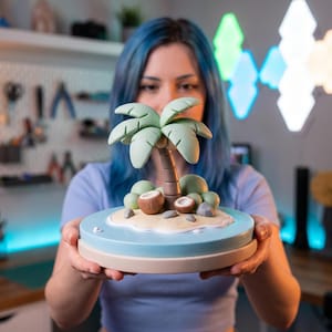 May include: A miniature tropical island sculpture featuring a central palm tree with light green fronds and a brown trunk. The island base includes sandy terrain, two halved brown coconuts, and small gray rocks, all set on a circular blue water platform.