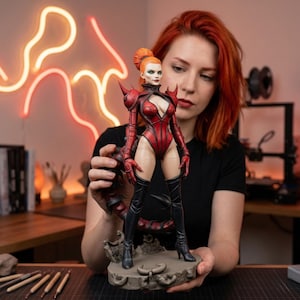 May include: A detailed figurine of a female character in a red bodysuit with black boots and spiked shoulder pads. The figure has orange hair and is standing on a gray base. The figurine is being held by a person with red hair.