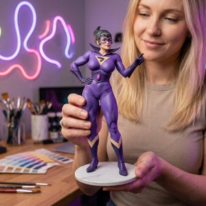 May include: A purple superhero figurine with a black mask, gloves, and boots. The figurine has a gold emblem on the chest and gold accents on the legs. It is displayed on a white marble-like base.