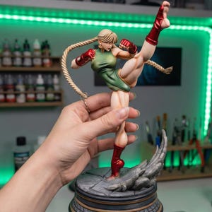 May include: A detailed figurine of a female character in a dynamic fighting pose. The figure wears a green leotard, red gloves, and boots. Her blonde hair is styled in two long braids. The base is gray and the figure is held by a hand.
