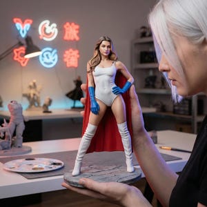 Può includere: Statuetta di una supereroina in body bianco, guanti blu e stivali bianchi, con mantello rosso. La statuetta è su una base circolare grigia. Lo sfondo include insegne al neon e altre sculture.