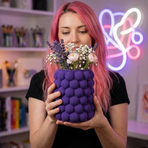 May include: A purple, textured vase filled with lavender, baby's breath, and pink flowers. The vase has a unique bubble-like design. The person holding the vase is wearing a black shirt.