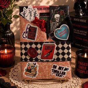 May include: A rectangular package wrapped in pink marble paper, featuring a black and white checkered ribbon, a red wax seal, and various stickers including a skull, a telephone, and text reading Thriller Book Club and In My Thriller Era.