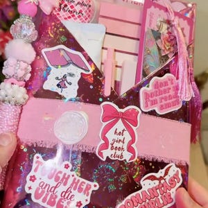 May include: A pink gift bag with a bow, decorative beads, and tassels. The bag features stickers with text like "Hot Girl Book Club" and "Romantasy Girlie." Inside are pink pens, a notebook, and other items.