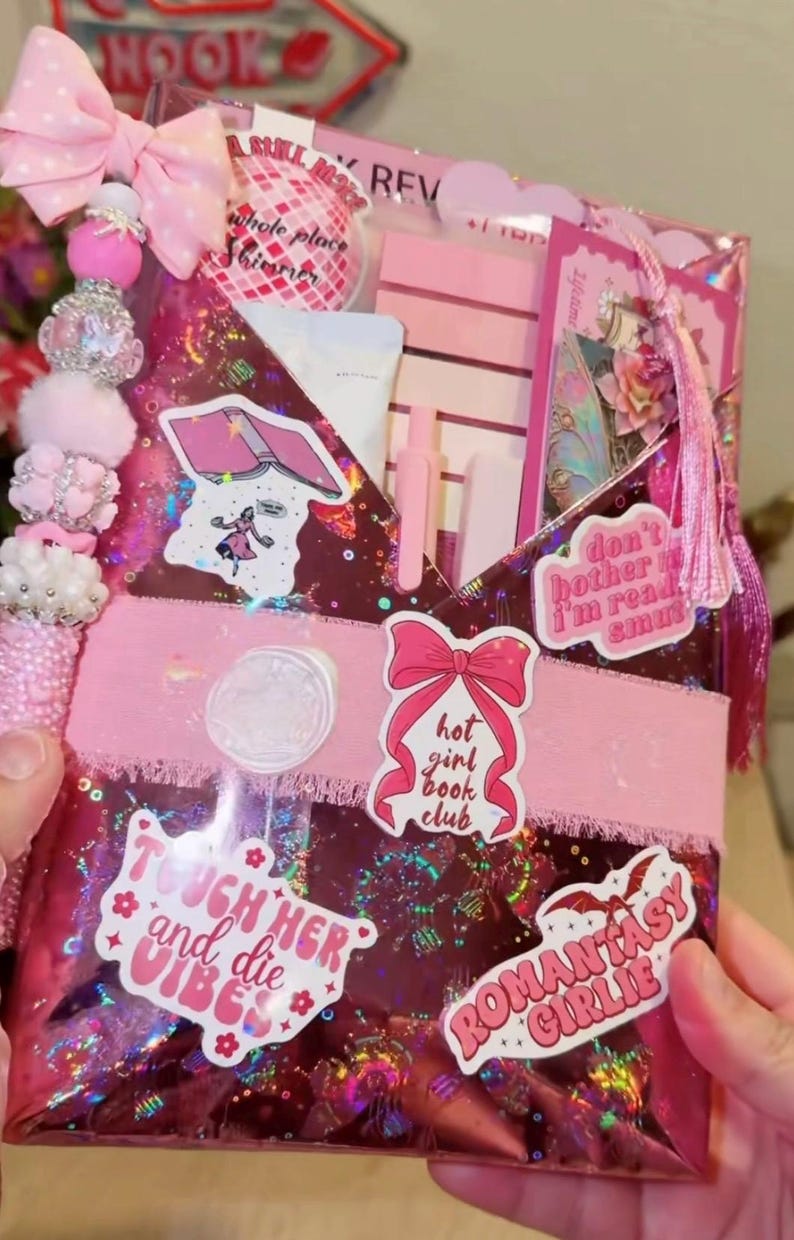 May include: A pink gift bag with a bow, decorative beads, and tassels. The bag features stickers with text like "Hot Girl Book Club" and "Romantasy Girlie." Inside are pink pens, a notebook, and other items.