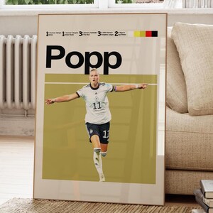 Op de afbeelding: Een ingelijste poster met de naam "Popp" in zwart en een foto van een voetballer in een wit en zwart uniform. De poster heeft een gele en witte achtergrond en een Duits vlagontwerp.