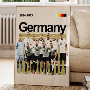 Op de afbeelding: Een ingelijste poster met het Duitse nationale voetbalteam. De poster toont het woord "Duitsland" in grote zwarte letters, met de jaren "2024-2025" erboven. De Duitse vlag staat in de rechterbovenhoek. Het team draagt witte en zwarte shirts.