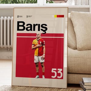 Pode incluir: Um poster emoldurado com um jogador de futebol com uma camisa vermelha e amarela. O poster tem o nome "Barış" em letras brancas grandes, um fundo vermelho e o número "53". O poster está encostado a uma parede numa sala de estar.