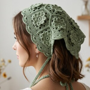 May include: A sage green crochet headscarf constructed from multiple hexagonal motifs, each featuring a raised central flower design. The headscarf has a scalloped edge and includes long, braided ties at the bottom for securing the accessory.