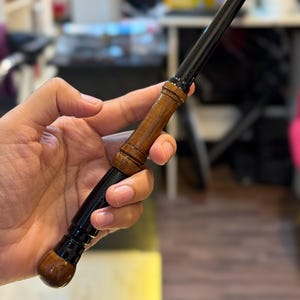 May include: A slender wand featuring a glossy black finish on the tapered tip and base, contrasted by a polished brown wooden handle with carved ring details. The handle has a rounded pommel at the end and a smooth transition to the black shaft.