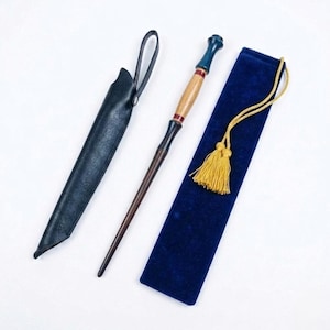 May include: A wooden wand with a dark brown handle and a light brown center, accented with red and blue details. It is accompanied by a black leather sheath and a blue velvet pouch with a gold tassel. The wand is a fantasy-themed item.