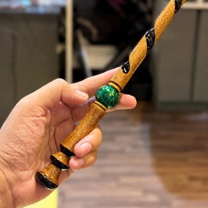 Puede incluir: Una varita de madera con un acabado de grano marrón natural y una incrustación negra en espiral a lo largo del eje. El mango incluye una cuenta esférica de color verde metálico situada entre la empuñadura y la sección superior.