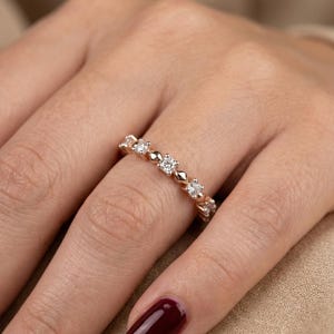 May include: A rose gold ring with a series of small, sparkling diamonds set in a repeating pattern. The ring is on a finger with a dark red nail polish. The ring's design includes diamond-shaped and round elements.