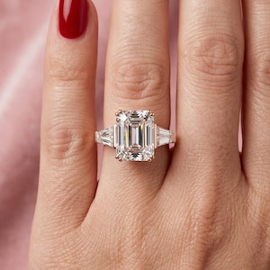 May include: A gold ring with a large, emerald-cut diamond center stone, flanked by two smaller, trapezoid-cut diamonds. The ring is set on a hand with a red nail polish.