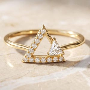 May include: A gold ring featuring a geometric triangle design. The frame is accented with small round pavé-set diamonds along two sides, while the third corner is highlighted by a single, larger triangular-cut diamond set within the gold structure.