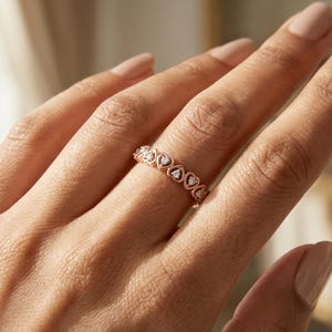 May include: A rose gold ring with a repeating heart design, each heart set with a small, clear stone. The ring is worn on a finger, highlighting its delicate and romantic style. The ring's design is symmetrical and elegant.