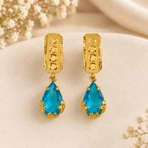 Könnte beinhalten: Ein Paar goldene Ohrhänger mit einem rechteckigen Oberteil mit vier kreisförmigen Vertiefungen und einem hängenden, tropfenförmigen, leuchtend blauen Edelstein in einer goldenen Fassung mit kleinen floralen Akzenten.