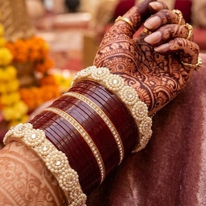 May include: Close-up of a hand adorned with henna and gold rings, showcasing a set of maroon bangles. The bangles are accented with gold bands and pearl-like embellishments, creating an elegant and traditional look.