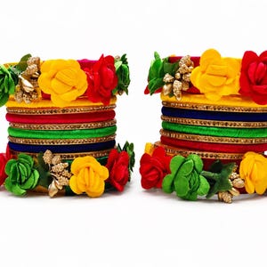 Könnte beinhalten: Zwei Stapel bunter Armreifen. Jeder Stapel zeigt Schichten aus roten, grünen, blauen und gelben Armreifen, verziert mit goldenen Verzierungen und leuchtenden Stoffblumen in Rot, Gelb und Grün.