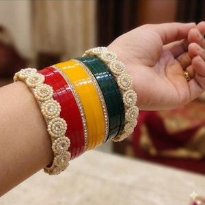 Könnte beinhalten: Ein Satz bunter Armreifen in Rot, Gelb und Grün, verziert mit einem funkelnden Strassband und perlenartigen Verzierungen. Die Armreifen sind auf einem Handgelenk gestapelt und zeigen das aufwendige Design und die leuchtenden Farben.
