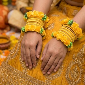 May include: Close-up of two hands adorned with multiple yellow bangles. Each bangle is decorated with yellow fabric roses and green accents. The hands rest on a yellow embroidered garment, suggesting a festive or ceremonial occasion.