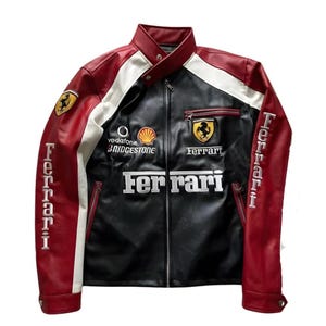 Op de afbeelding: Een zwarte, rode en witte Ferrari racejas. De jas heeft het Ferrari-logo, sponsorlogo's en het woord "Ferrari" in witte letters. De mouwen zijn rood met witte letters. De jas heeft een ritssluiting.