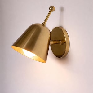 May include: A brass wall sconce with a conical shade and a round backplate. The sconce has a small, adjustable arm and a warm, inviting light emanating from the shade. The fixture is mounted on a white wall.