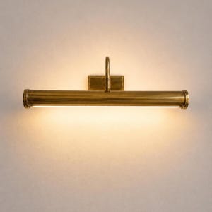 May include: A brass-toned picture light fixture with a rectangular backplate and a curved arm. The horizontal light has a cylindrical design with a bright, linear light source. The fixture is mounted on a neutral-colored wall.