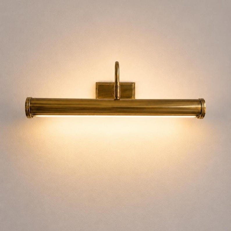 May include: A brass-toned picture light fixture with a rectangular backplate and a curved arm. The horizontal light has a cylindrical design with a bright, linear light source. The fixture is mounted on a neutral-colored wall.