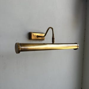 May include: A vintage-style brass picture light fixture. The fixture has a rectangular wall mount, a curved arm, and a cylindrical light housing. The brass finish gives it a classic, elegant look. The light is designed to illuminate artwork.