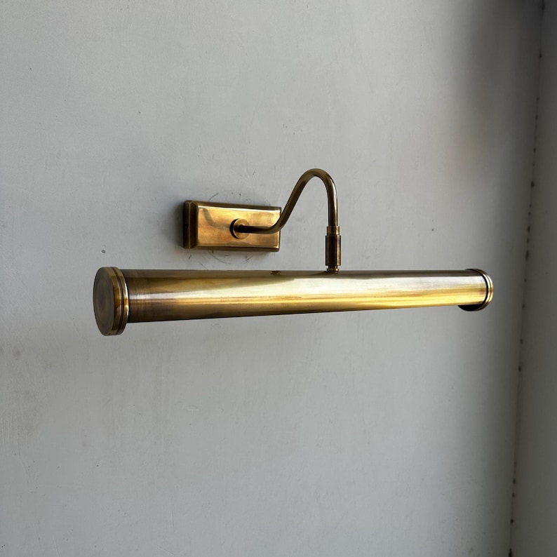 May include: A vintage-style brass picture light fixture. The fixture has a rectangular wall mount, a curved arm, and a cylindrical light housing. The brass finish gives it a classic, elegant look. The light is designed to illuminate artwork.