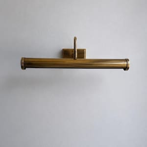 Peut inclure: Un éclairage de tableau en laiton, avec une plaque arrière rectangulaire et un bras incurvé. La lampe a un abat-jour cylindrique, conçu pour être monté au mur. Le luminaire est en laiton, avec une teinte dorée chaude. Le design est classique.